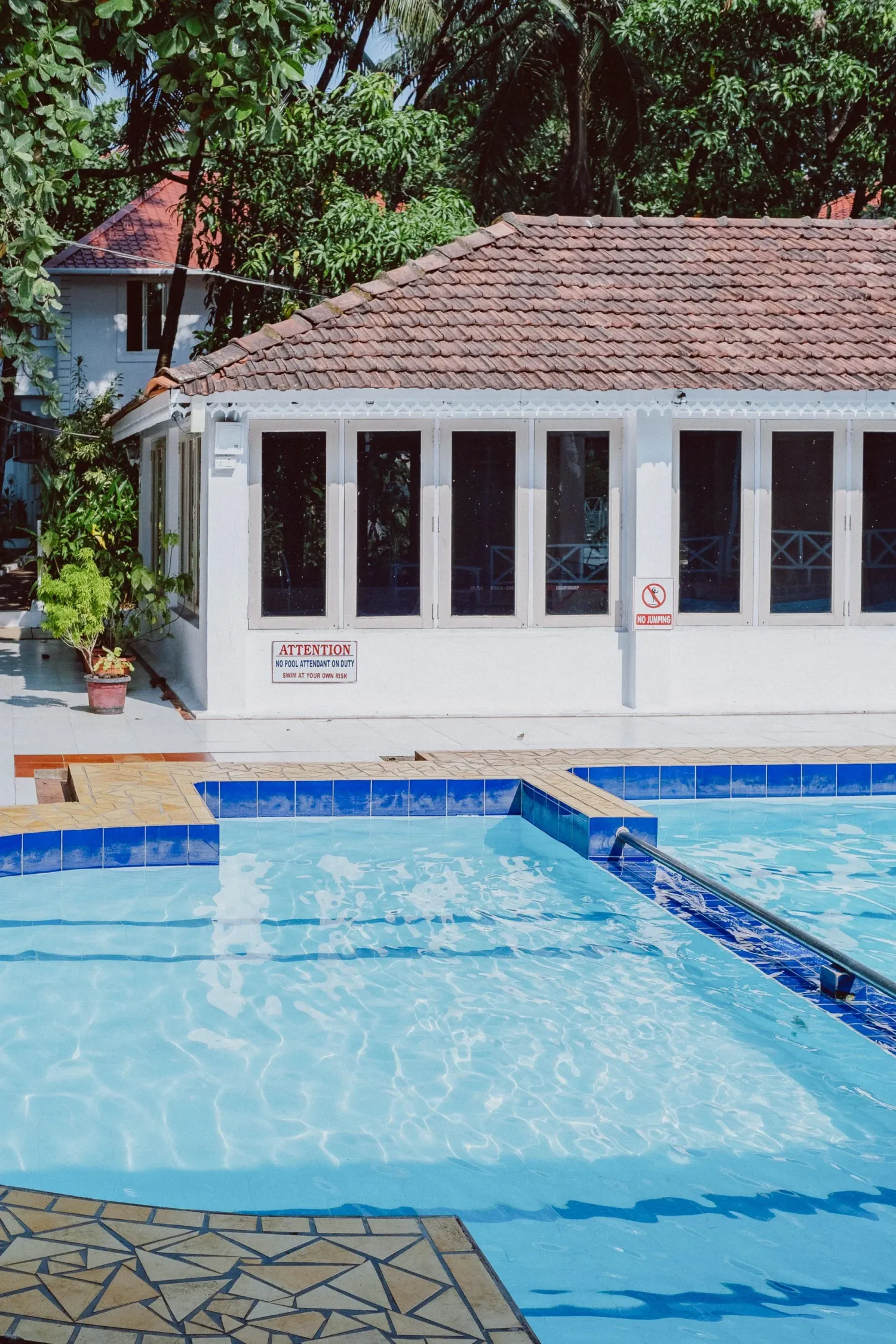 A bright blue outdoor swimming pool beside a white colonial-style poolhouse surrounded by tropical trees.