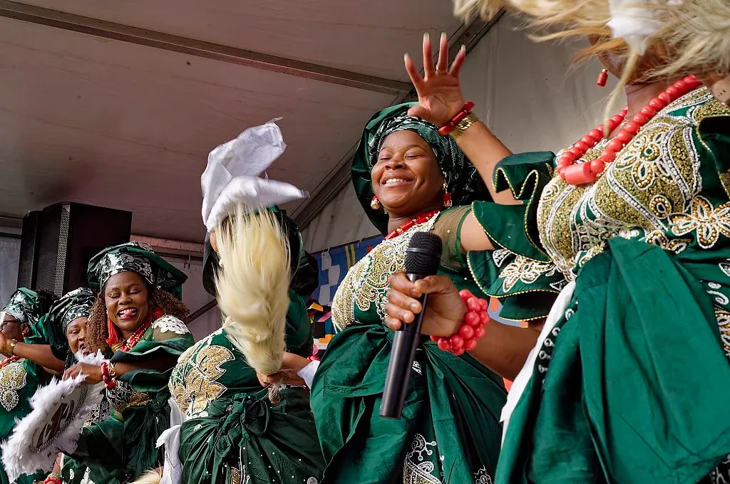 Women in traditional green and gold Nigerian attire perform and celebrate joyfully on stage at a cultural event.
