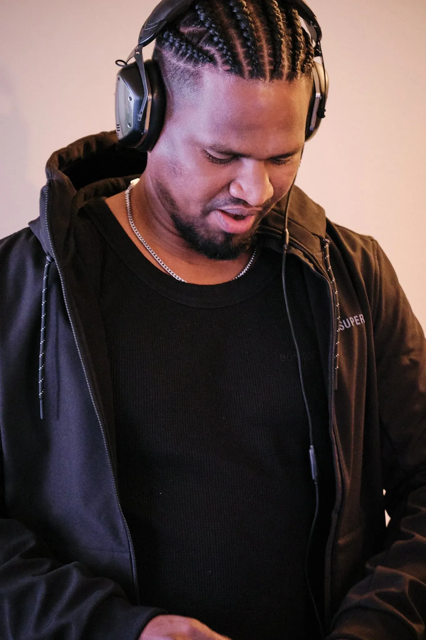 Male DJ with cornrow braids wearing black outfit and headphones, focused on mixing music.