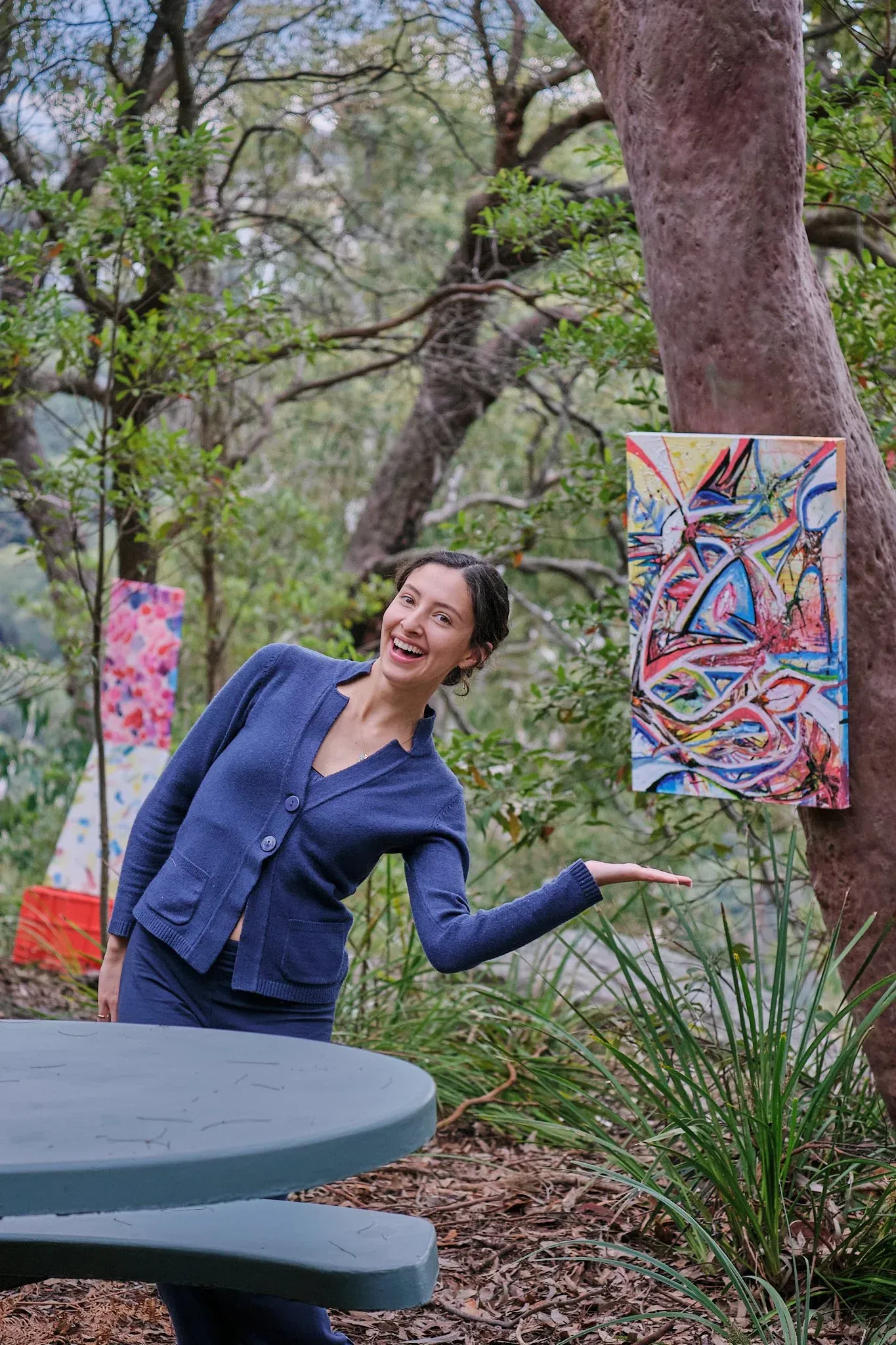 Laughing woman in blue cardigan gesturing toward vibrant abstract painting on tree.