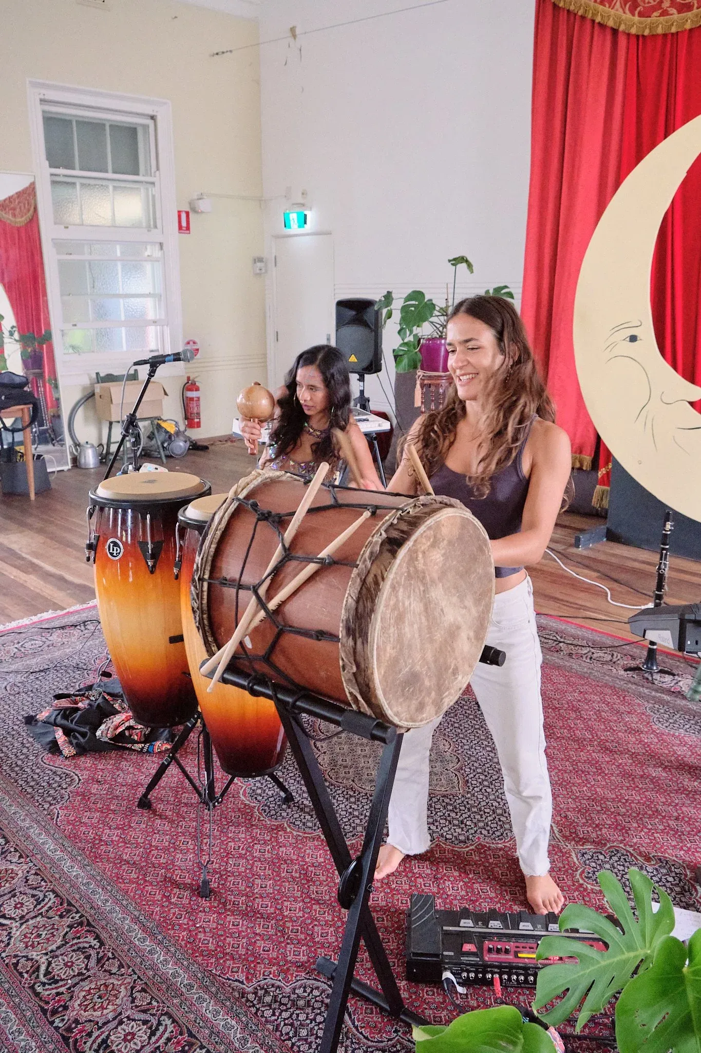 Two women play large drums including a taiko and conga at an indoor world music performance.