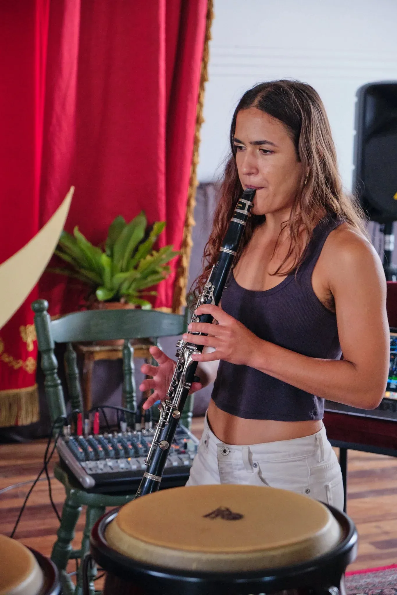 Female musician plays clarinet beside bongo drums in a bohemian studio with red curtains.