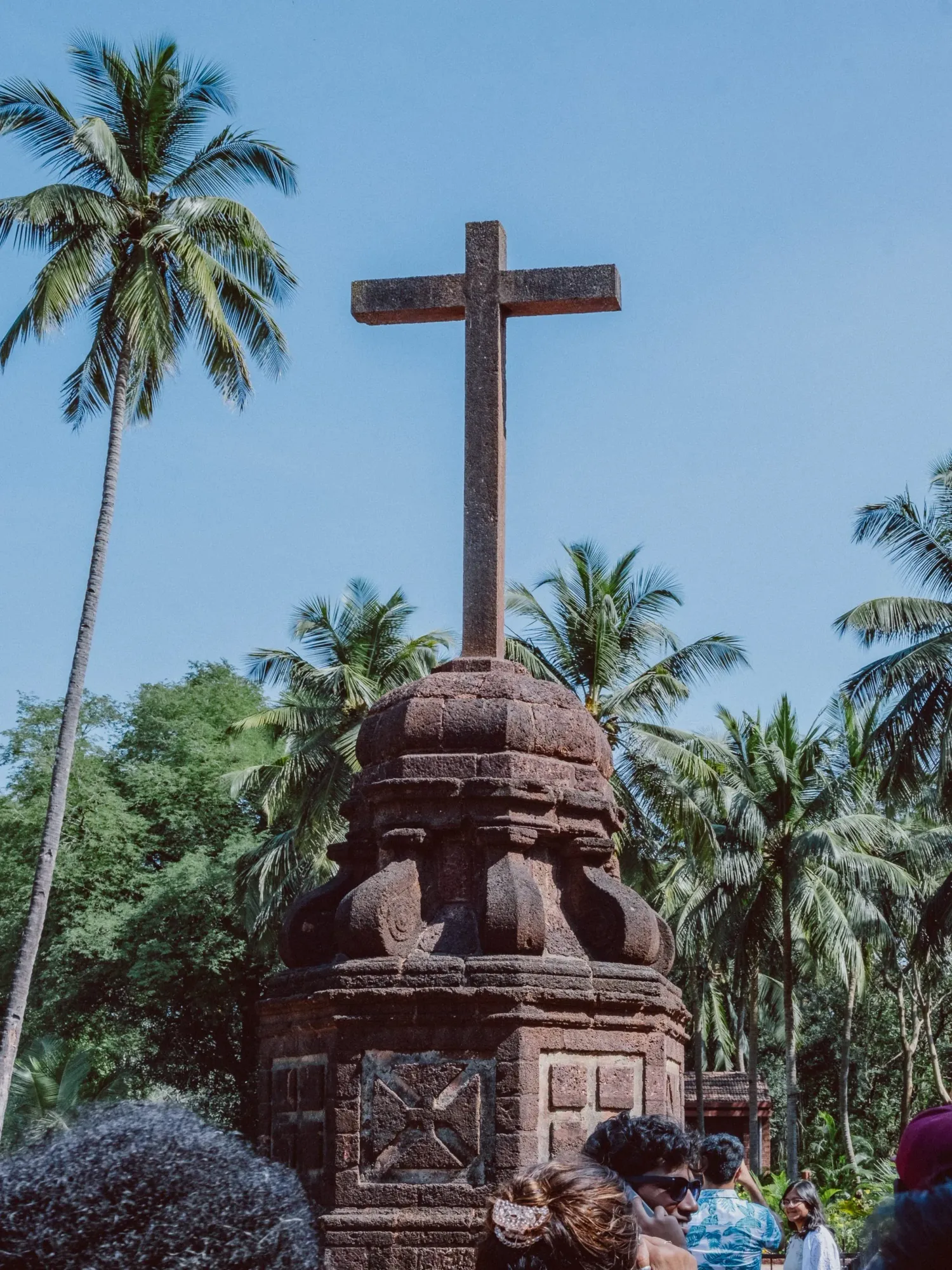 Ancient stone cross monument surrounded by coconut palm trees against clear blue sky.
