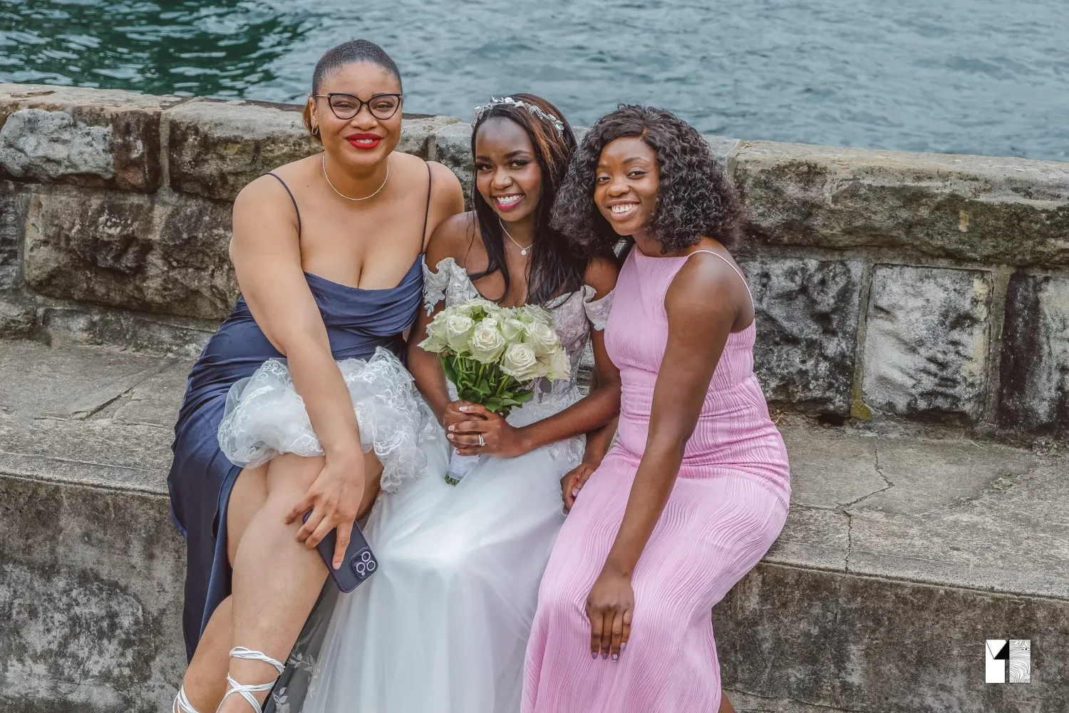 Bride with two bridesmaids in blue and pink dresses by stone wall waterfront.