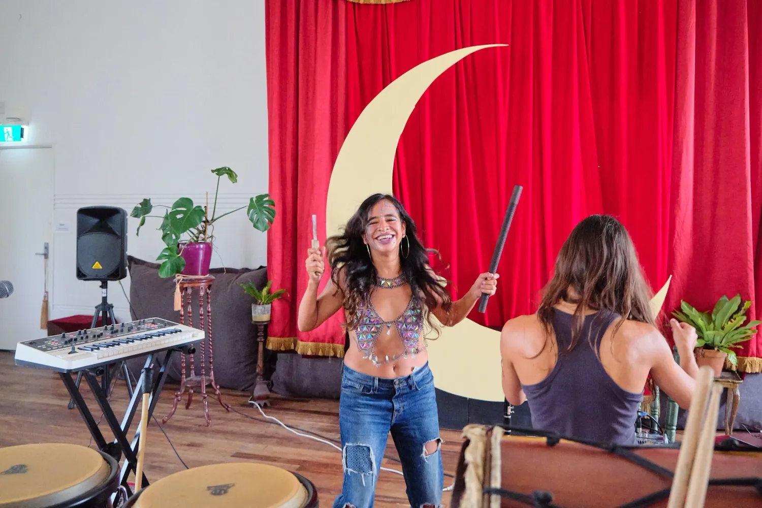 Two women perform music in a bohemian studio with red curtains and a crescent moon backdrop.