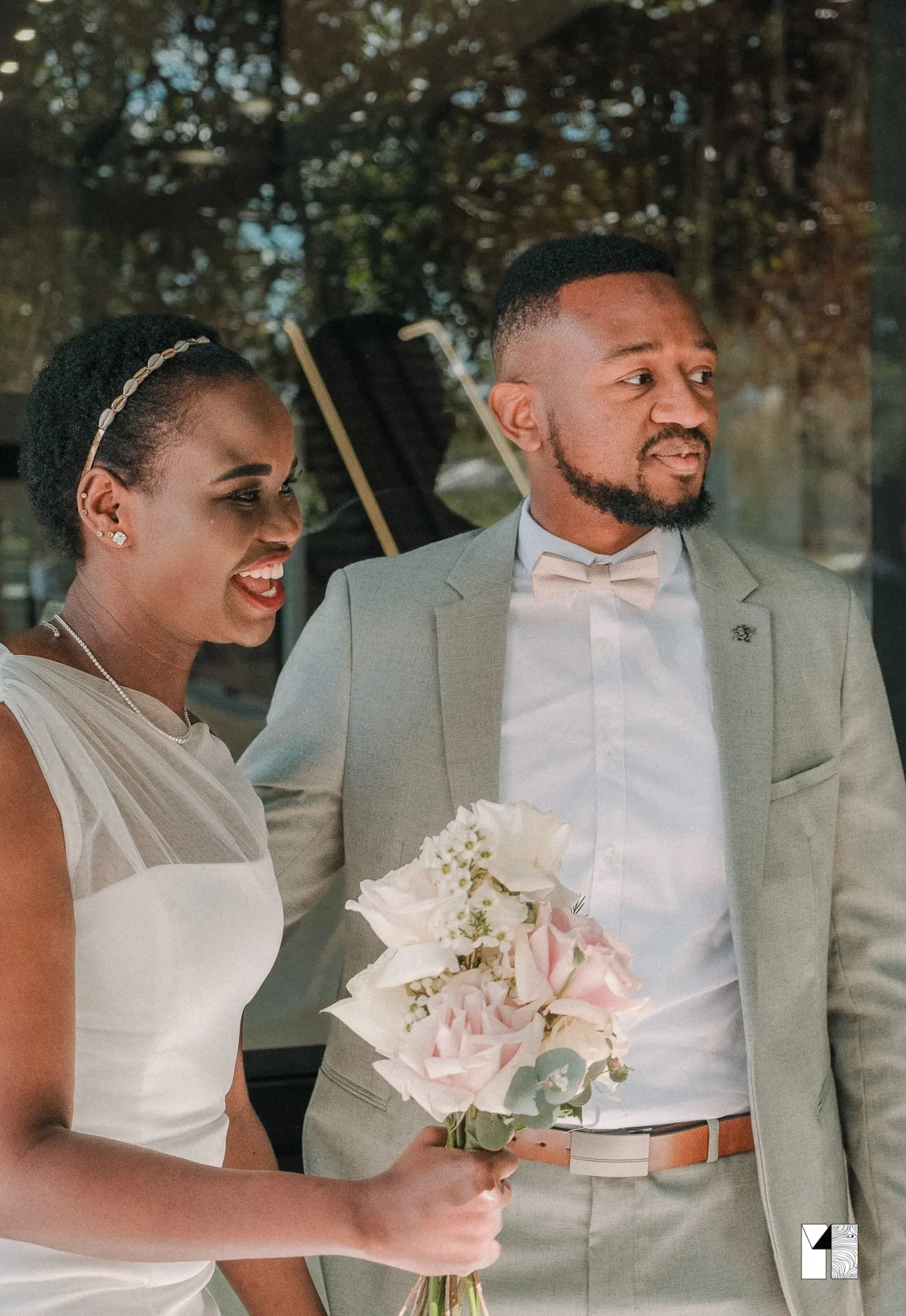 Bride and groom standing together, groom in gray suit with bow tie, bride holding pink bouquet.