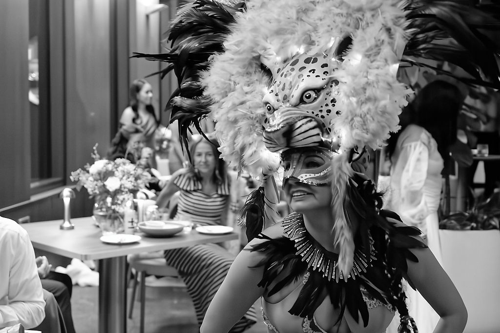 Black and white photo of a woman in a jaguar headdress with feathers and beaded necklace at an outdoor event with guests behind.