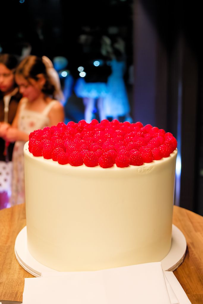 Wedding cake with smooth white frosting topped with fresh raspberries on a wooden table, guests blurred in the background.