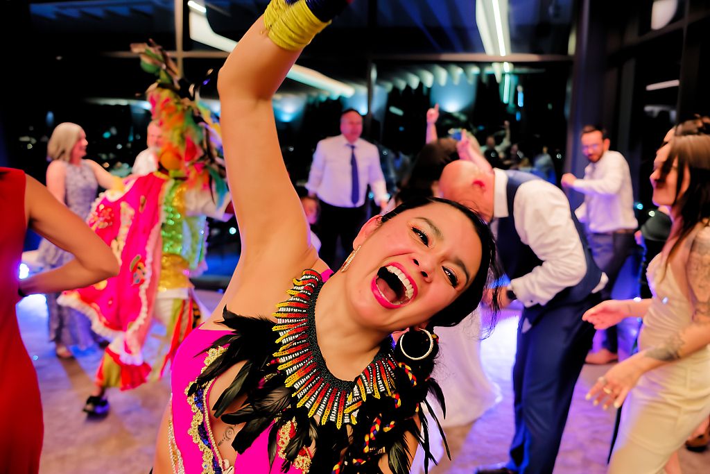Woman in a feathered tribal-style costume and beaded collar laughs joyfully with her arm raised while dancing at a lively party.