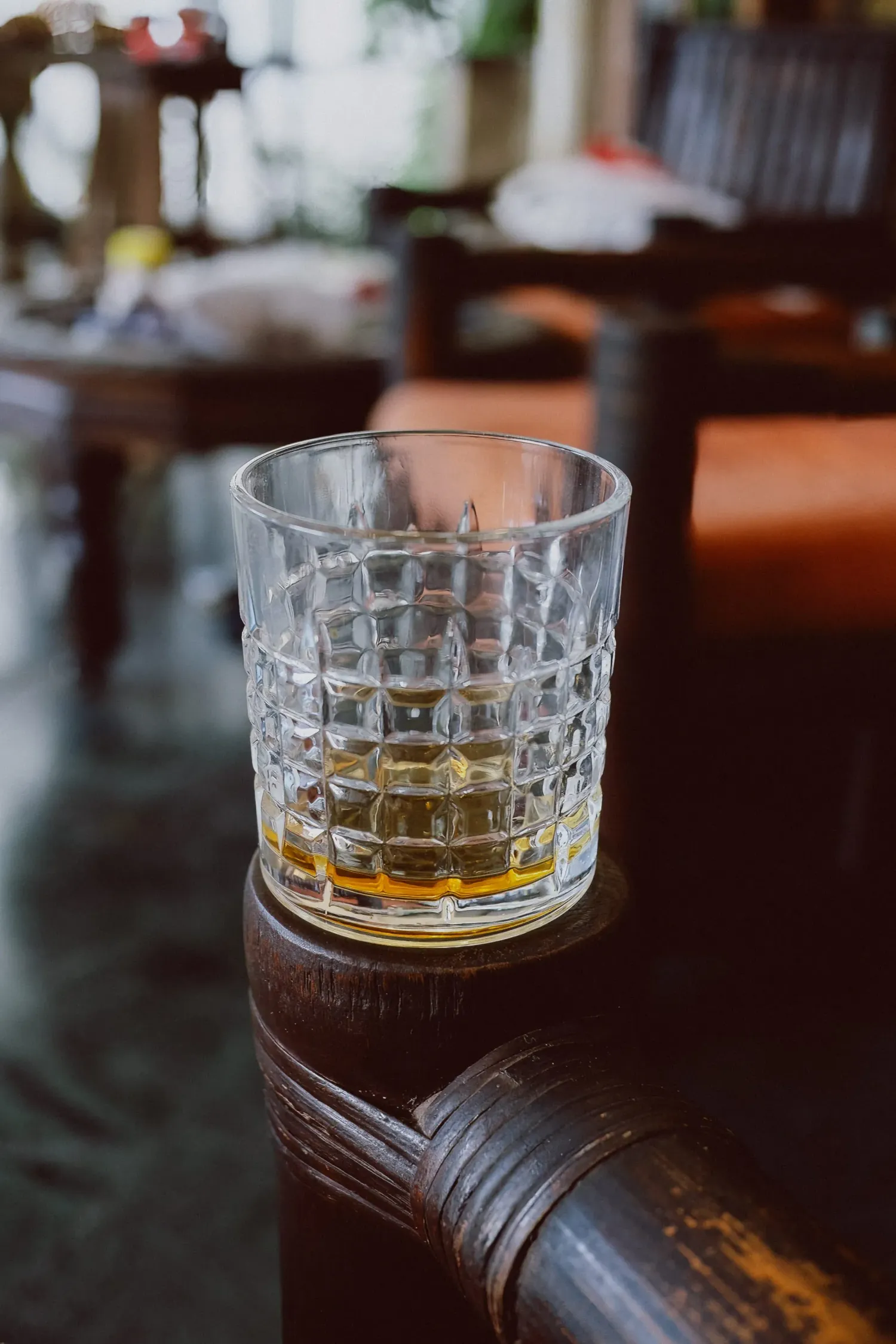 Crystal cut glass of whiskey resting on dark wooden armchair in blurred bar interior.