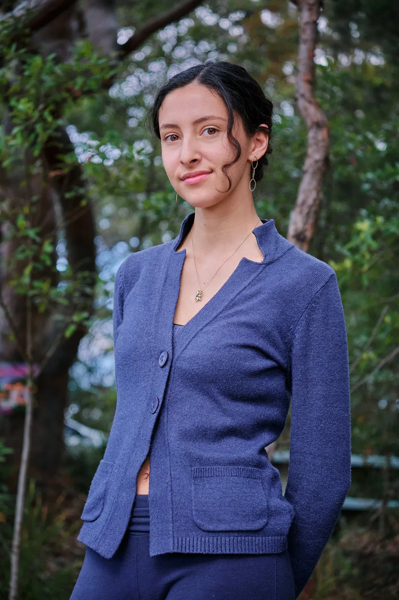 Smiling woman in blue cardigan standing outdoors among trees wearing a delicate necklace.