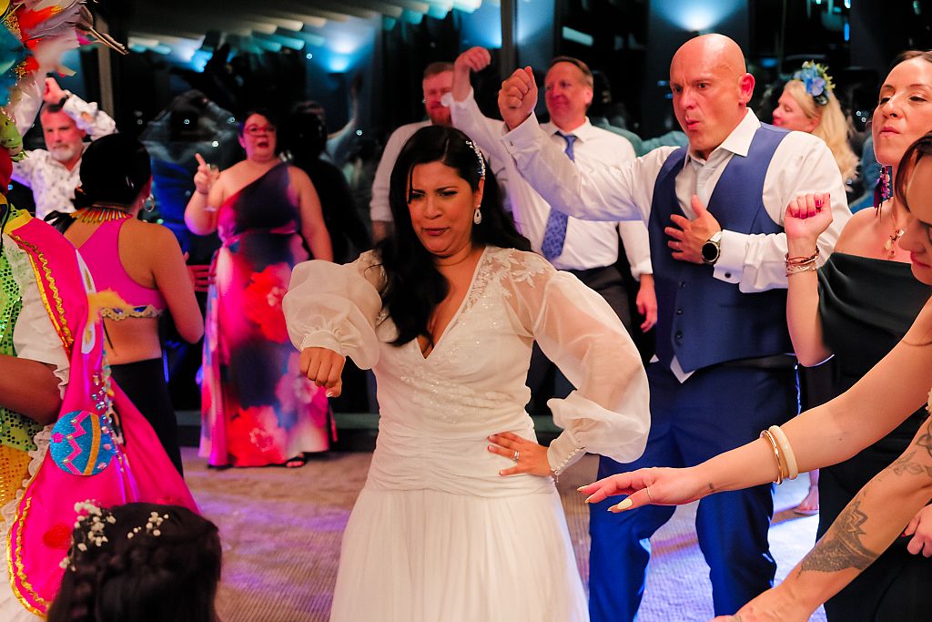 Bride in a white dress dances joyfully at a lively wedding reception surrounded by cheering guests in colorful formal attire.