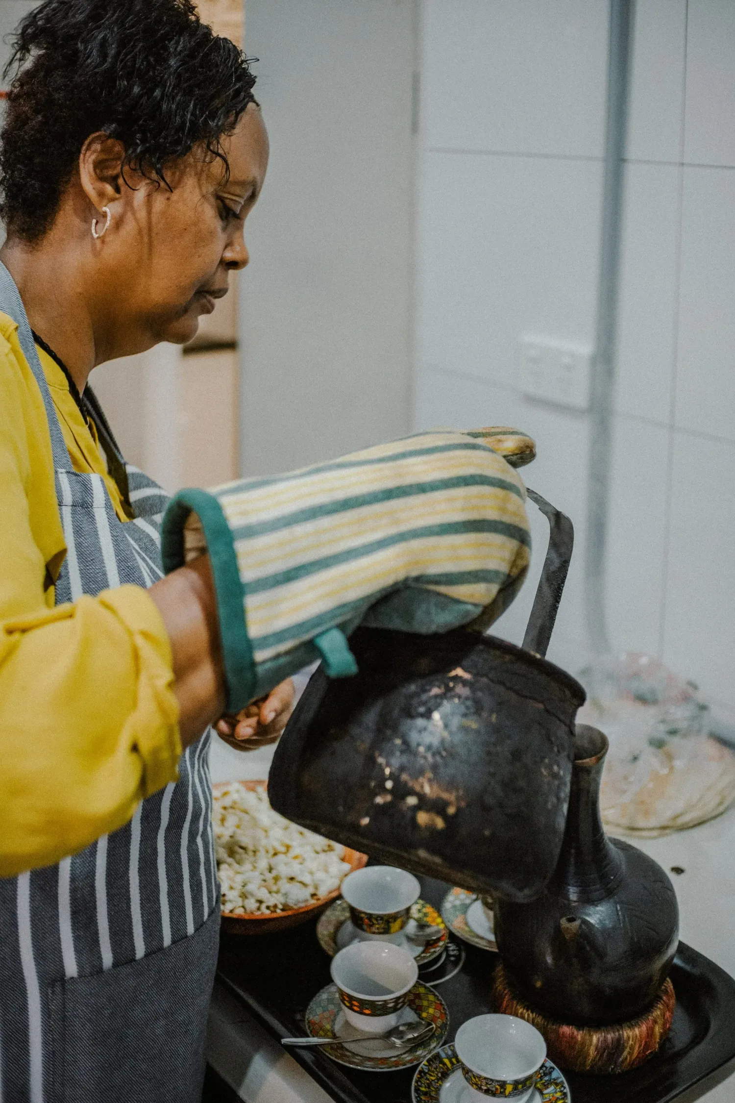 Woman in apron and oven mitt pouring from a traditional black Ethiopian coffee pot on a stove.