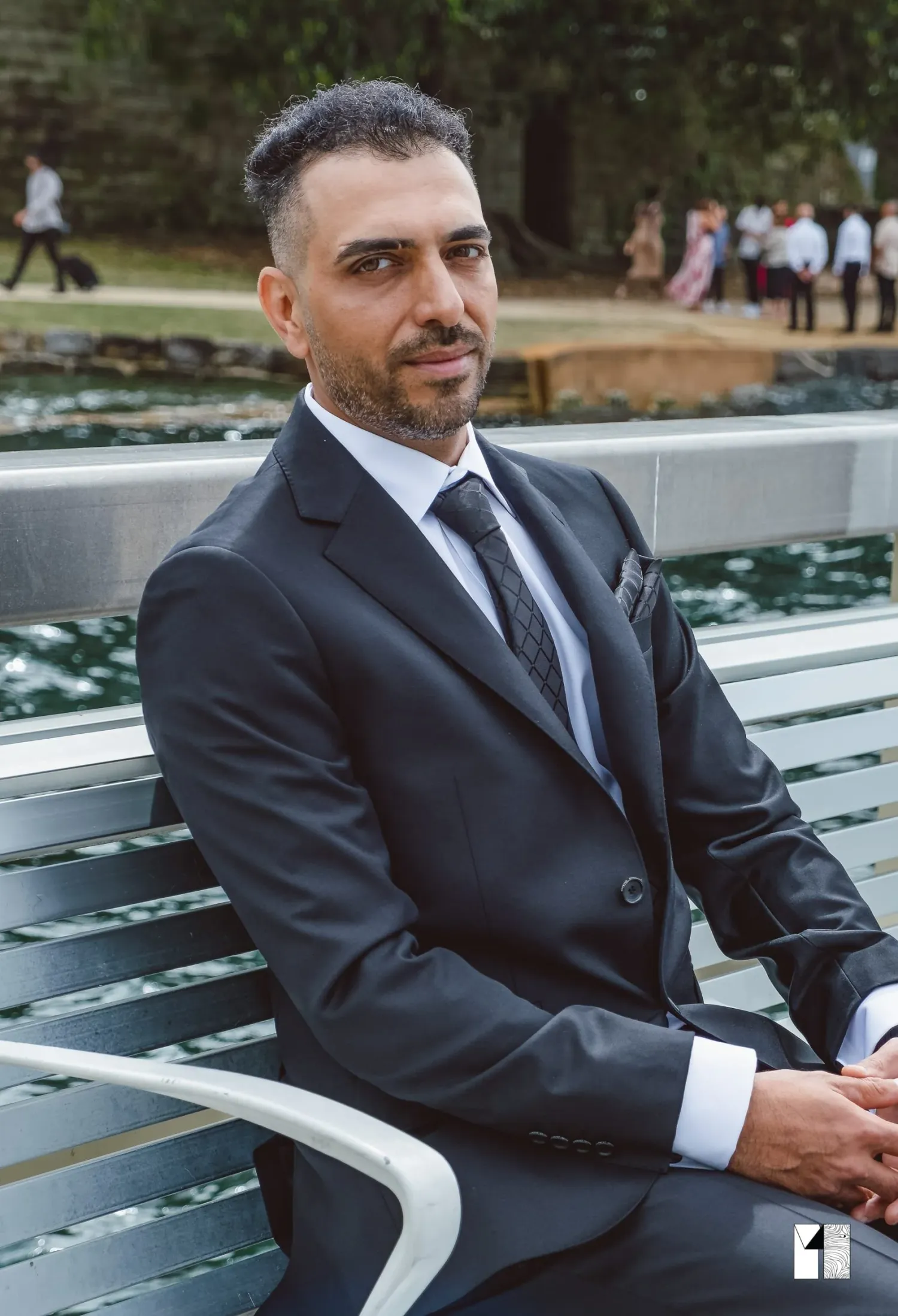 Groom in dark suit sitting on bench near waterfront on wedding day.