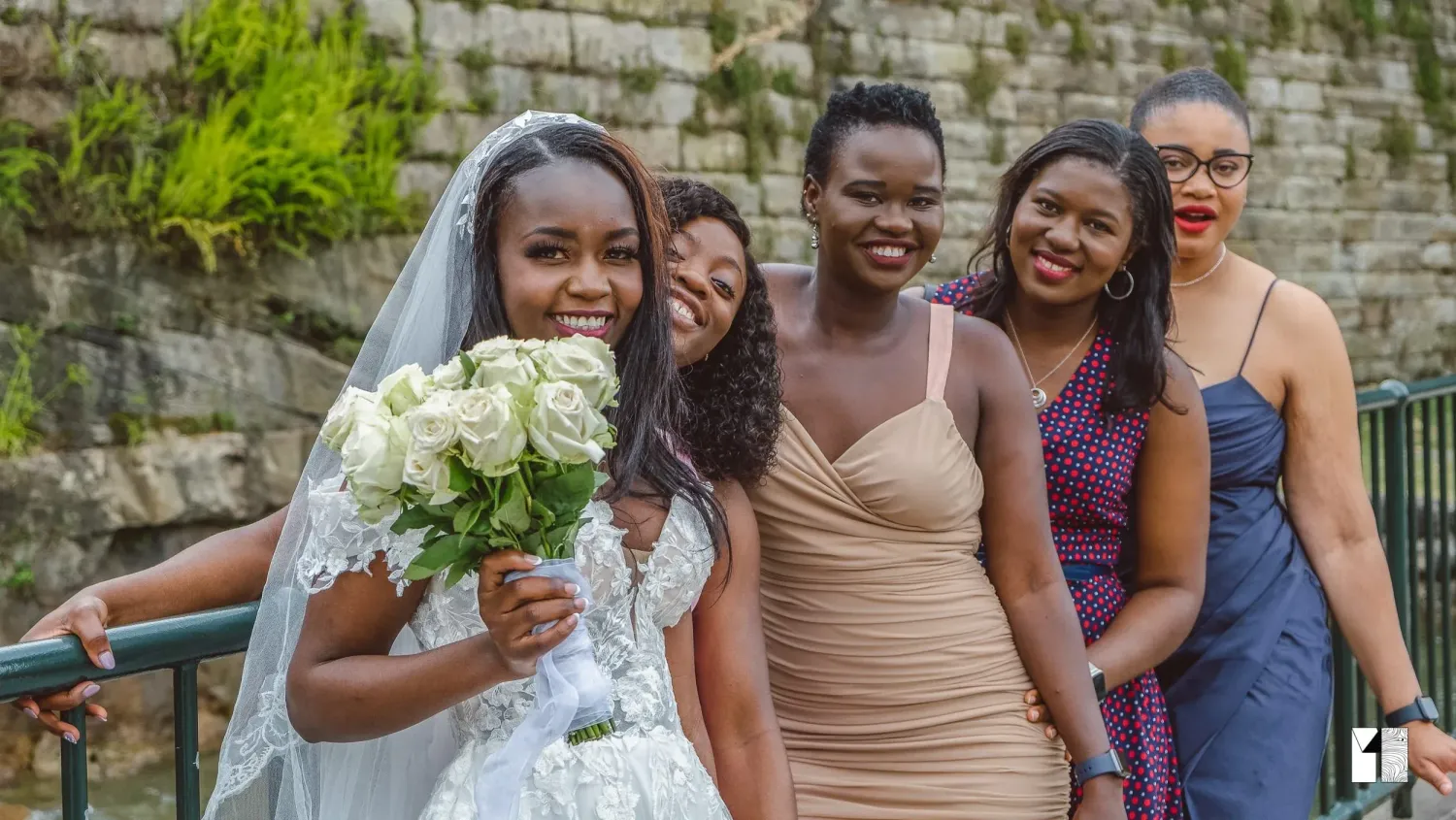 Radiant bride with bouquet poses with smiling bridesmaids in colorful dresses outdoors.