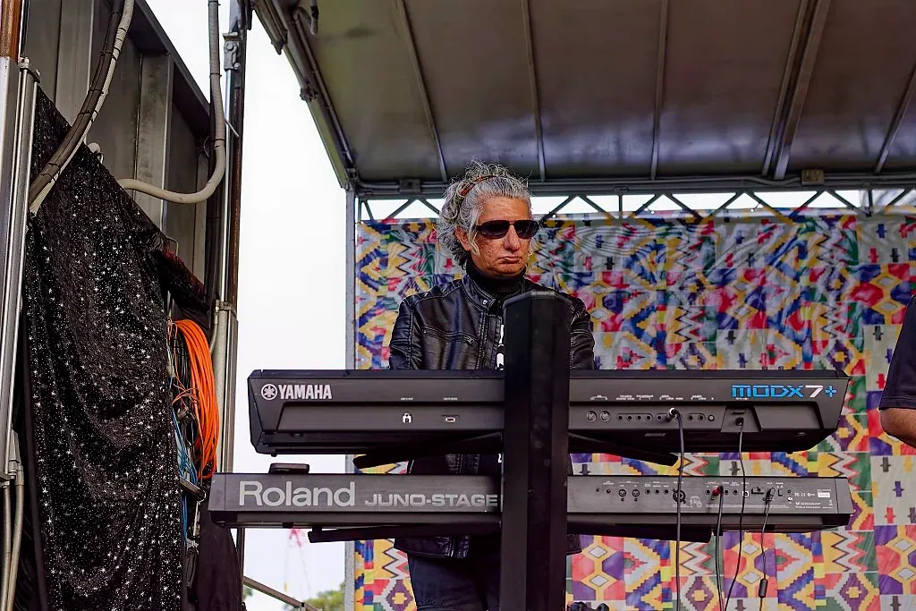 A musician plays a Roland keyboard on stage wearing sunglasses at an outdoor music festival.