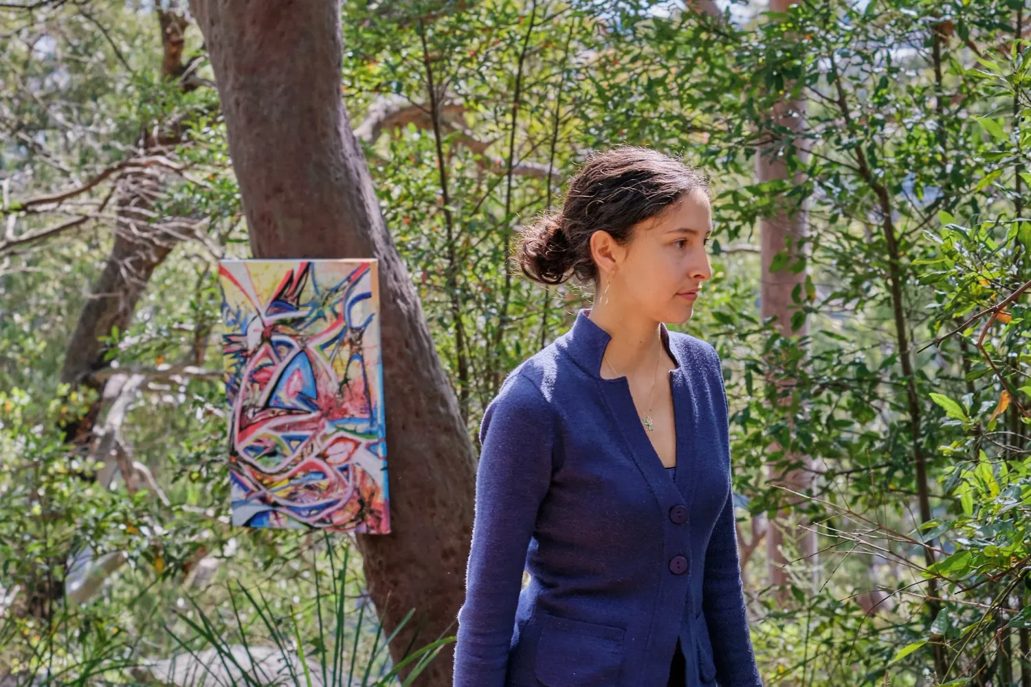 Woman in blue dress walking through forest beside colorful abstract artwork on tree.
