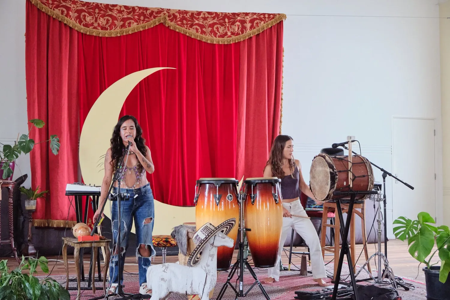 Two female musicians performing on stage with conga drums, red curtain and crescent moon decoration backdrop.