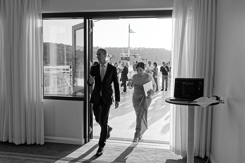 Black and white photo of the groom and bride walking through a glass door into the wedding venue, guests behind them.