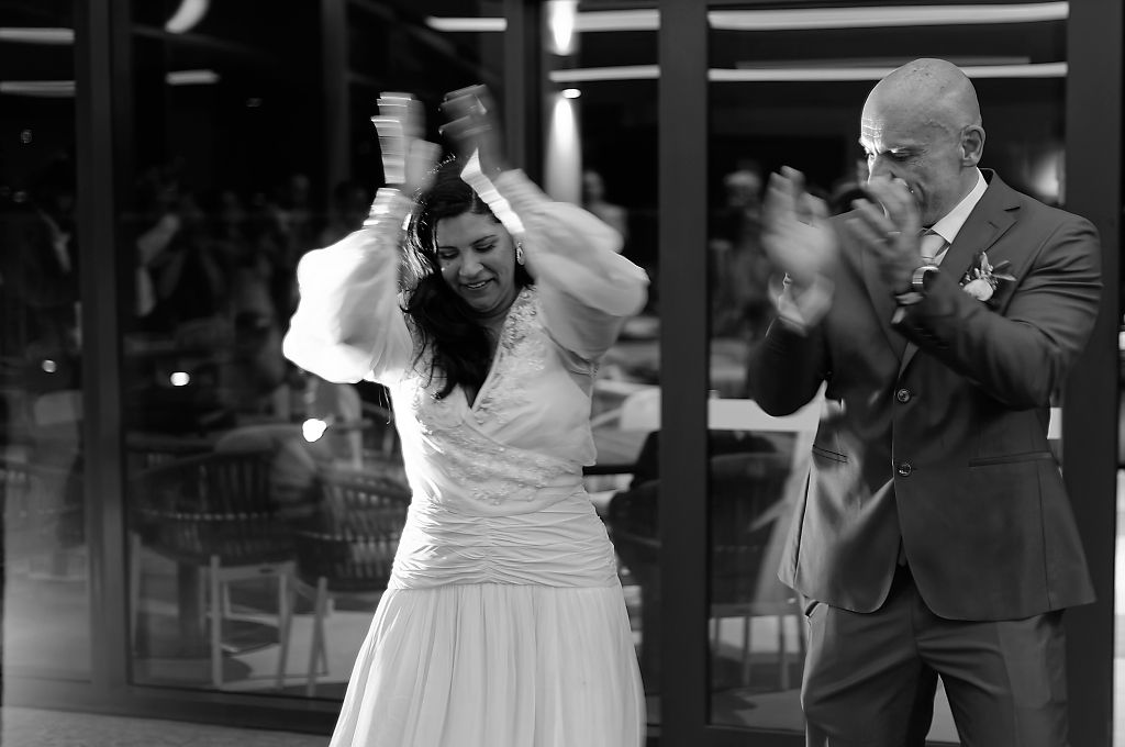 Black and white photo of a bride in a white dress and a groom in a suit clapping and dancing joyfully at their wedding reception.