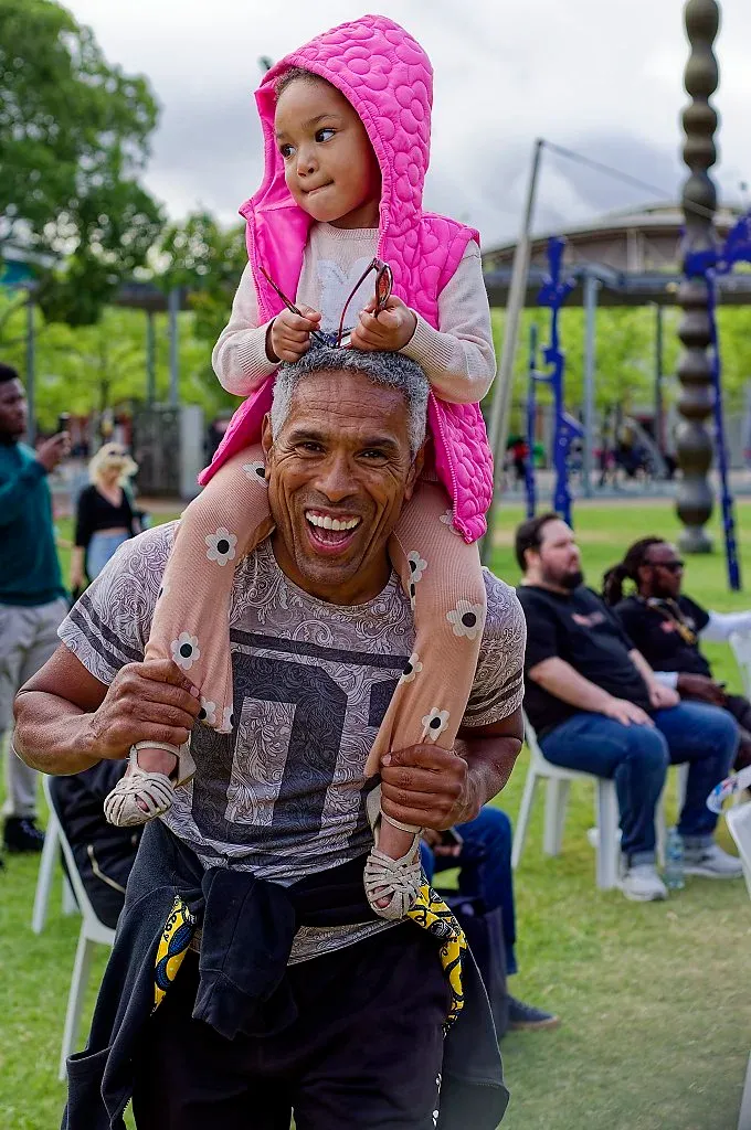 A smiling man carries a young child in a pink hoodie on his shoulders at an outdoor festival.