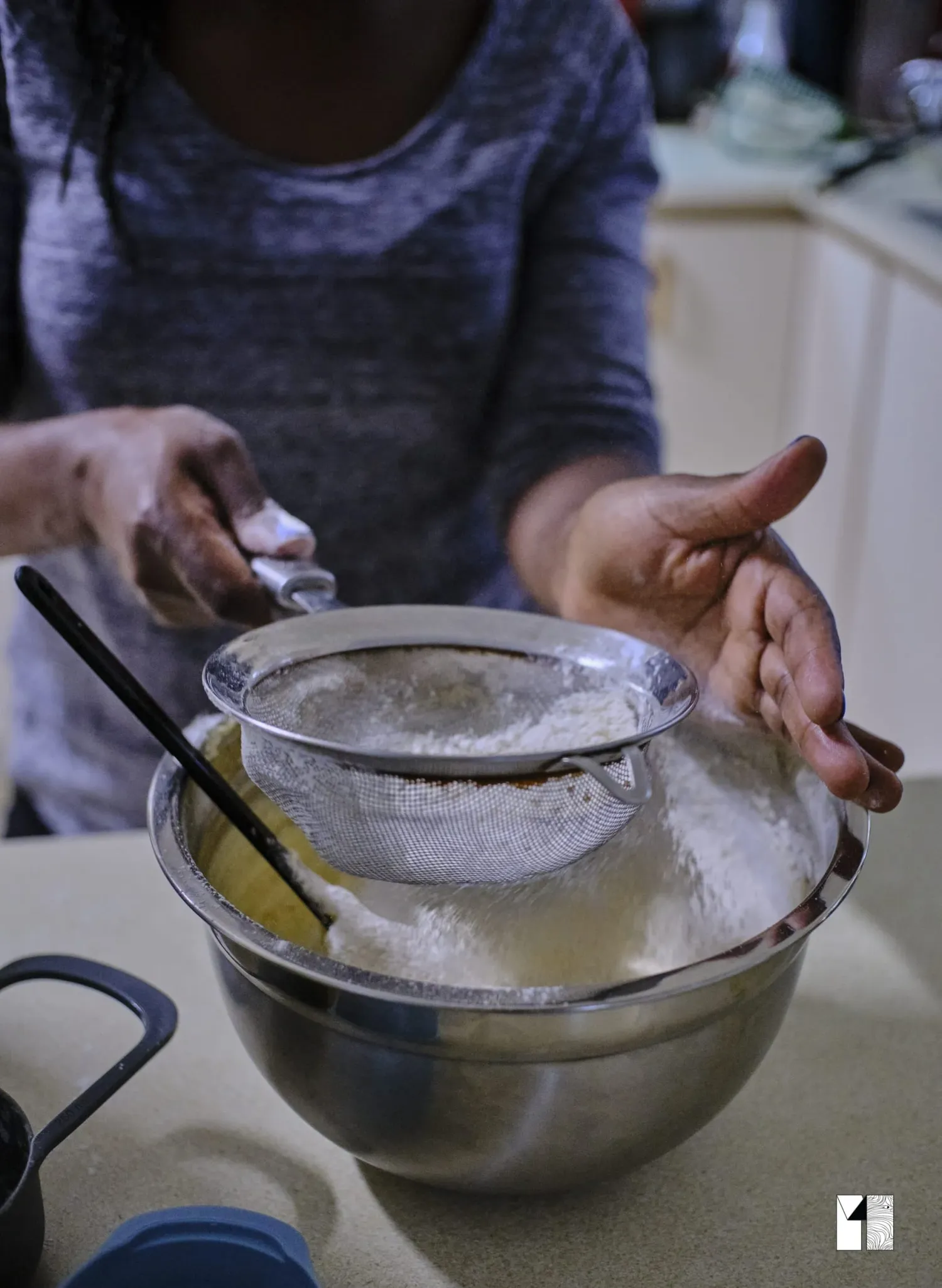 Person sifting flour through a mesh strainer into a stainless steel mixing bowl while baking.