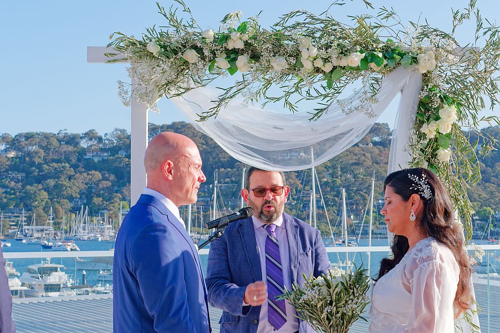 Outdoor wedding ceremony under a floral arch with a bride holding a bouquet facing the groom and officiant, with a marina and hills behind.