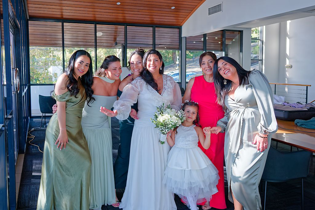 A bride in a lace gown holding a bouquet poses with smiling bridesmaids in green and red dresses and a flower girl in a bright room.