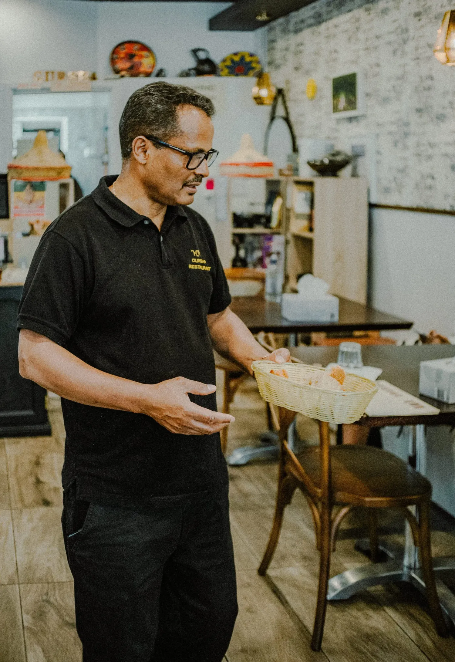 Restaurant staff member in black uniform presenting a basket of bread in a casual eatery.