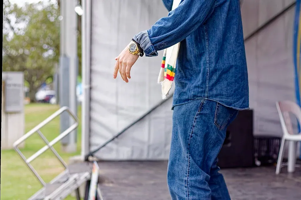 Person in denim outfit with colorful bracelet and gold watch performing on outdoor stage.