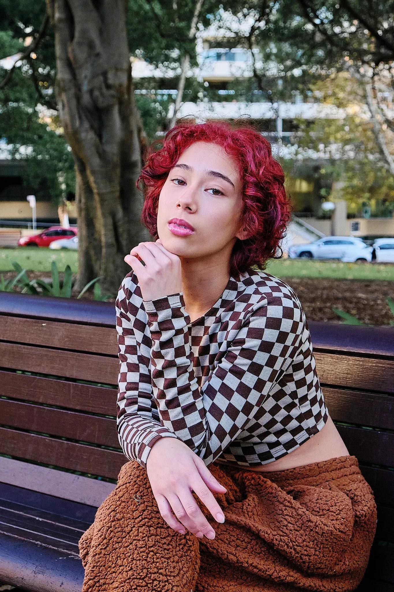 Young woman with red curly hair in checkered top sitting pensively on wooden park bench.