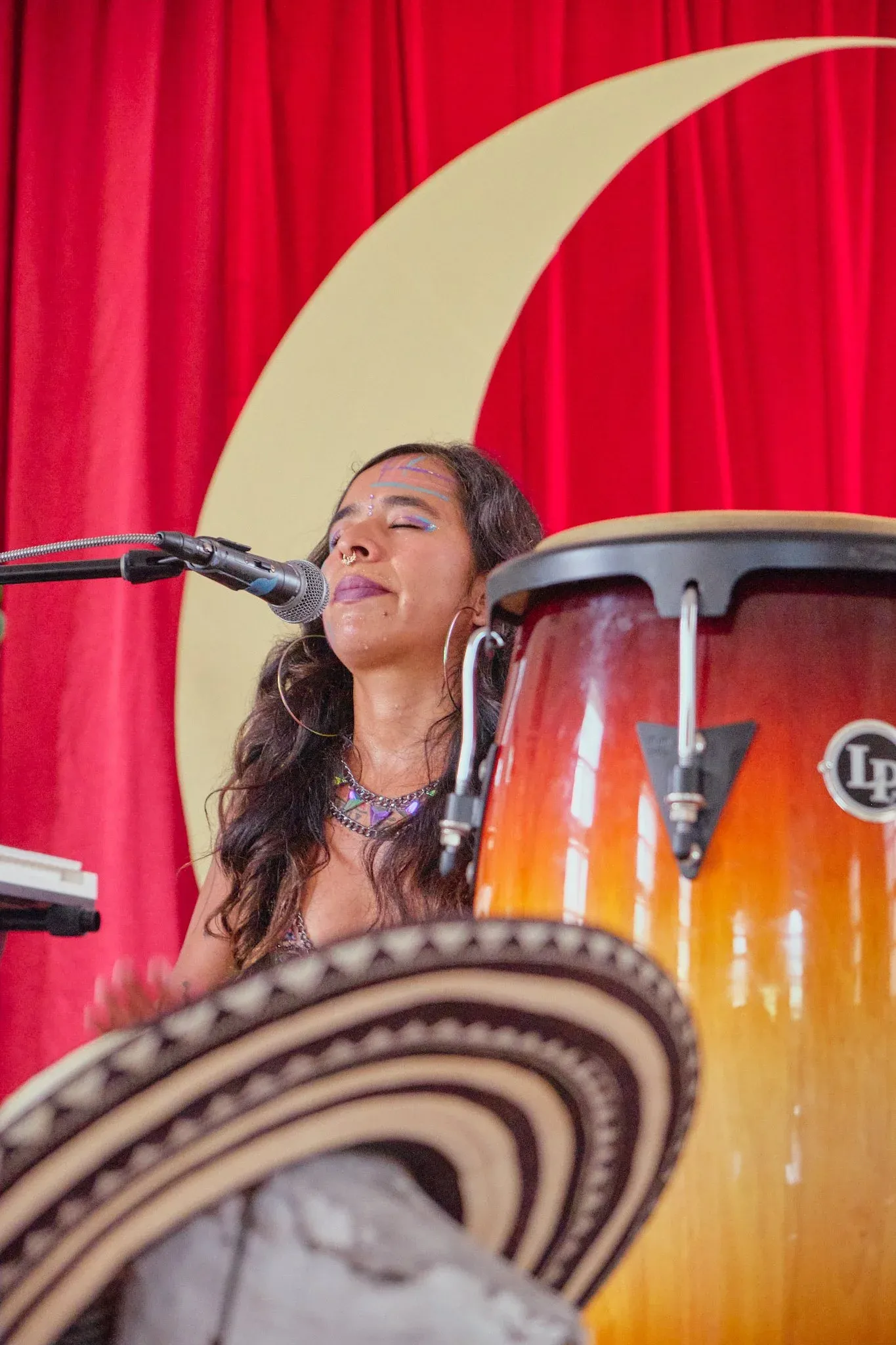 Female musician singing at microphone beside LP conga drums with red curtain and crescent moon backdrop.