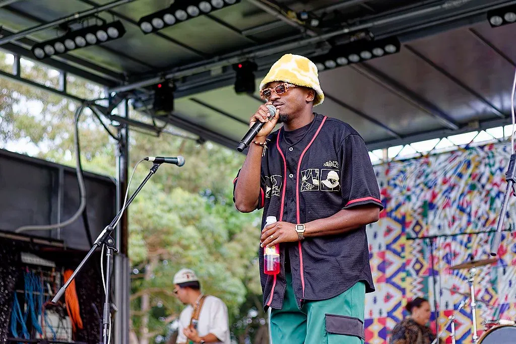 A young performer in a yellow bucket hat sings into a microphone on an outdoor festival stage.