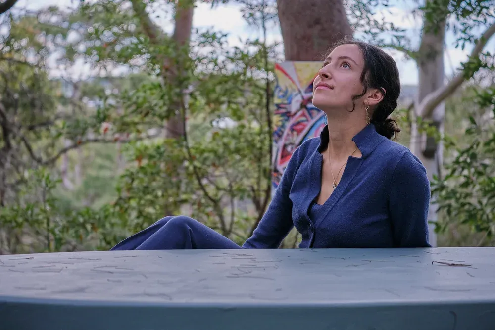 Woman in blue suit sitting at outdoor table, looking up thoughtfully surrounded by trees.