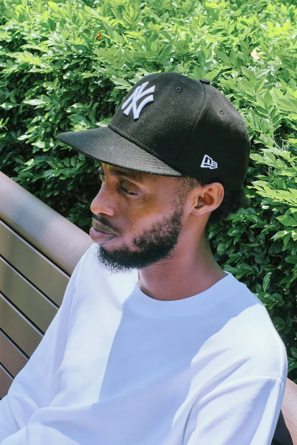 Side profile of Black man wearing black NY Yankees New Era cap seated near green hedges outdoors.