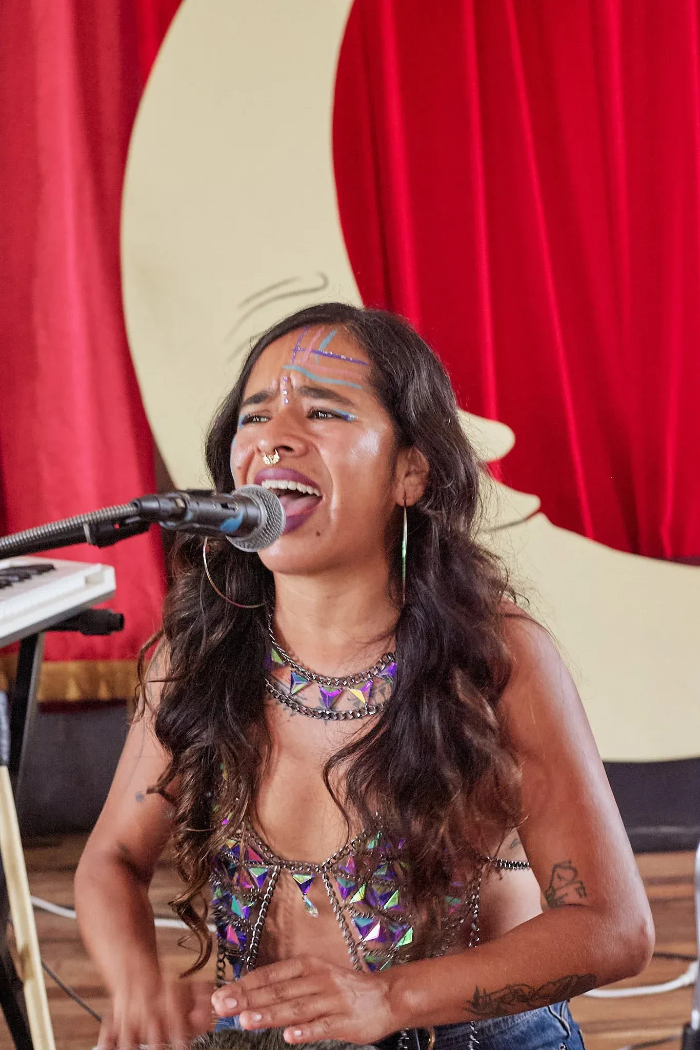 Female singer performing passionately at microphone with colorful face gems and jeweled costume on stage.