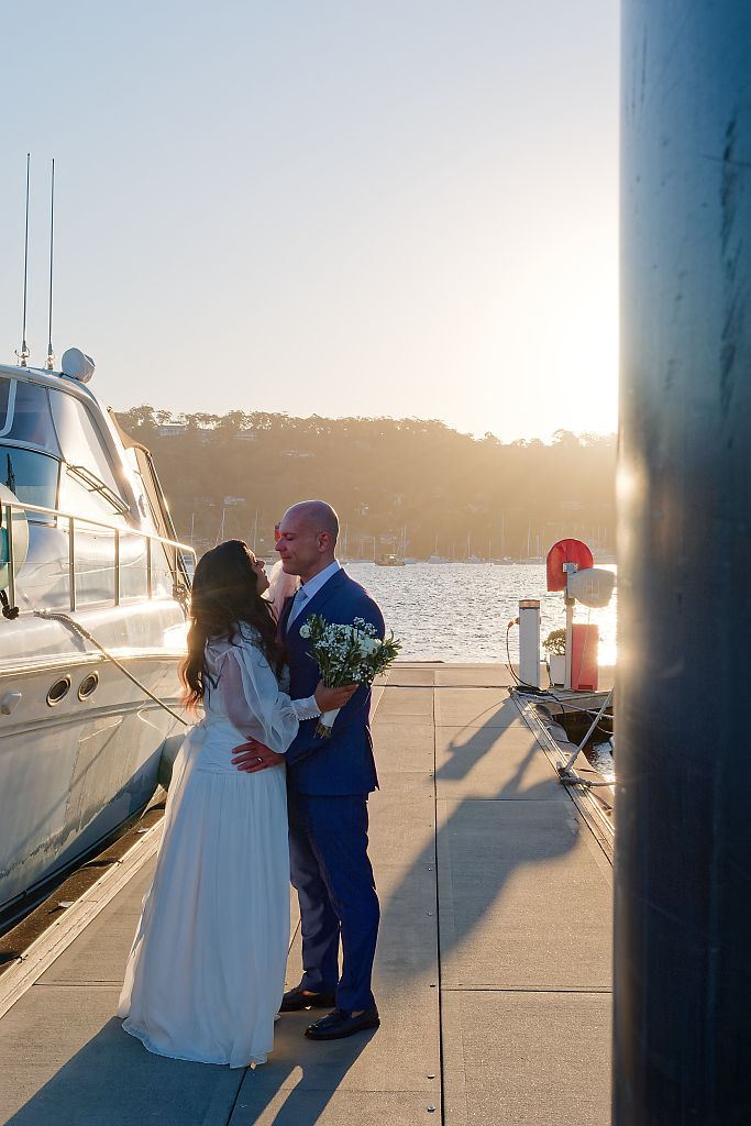 Bride and groom share an embrace on a marina dock at sunset beside a yacht, holding a white floral bouquet.