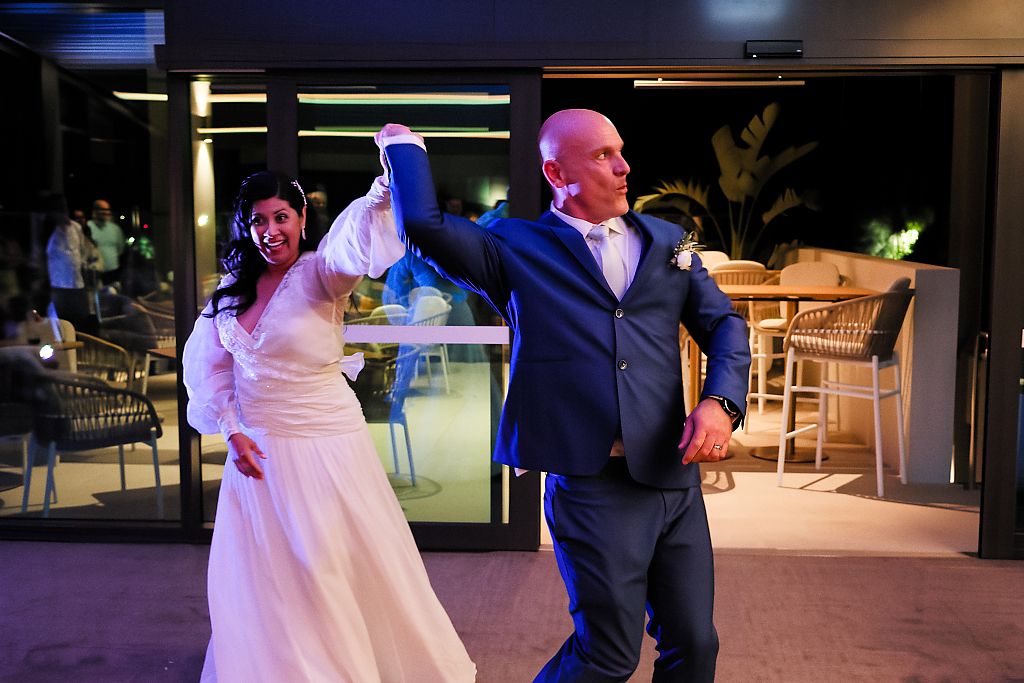 Bride in a white lace gown and groom in a navy suit dance joyfully with raised arms at an evening wedding reception.