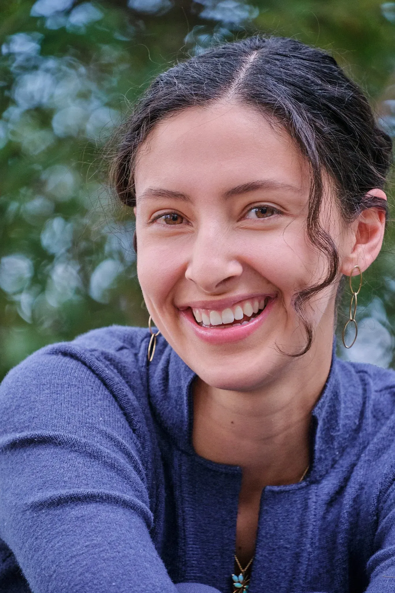 Close-up portrait of a smiling young woman with hoop earrings outdoors in a green natural setting.
