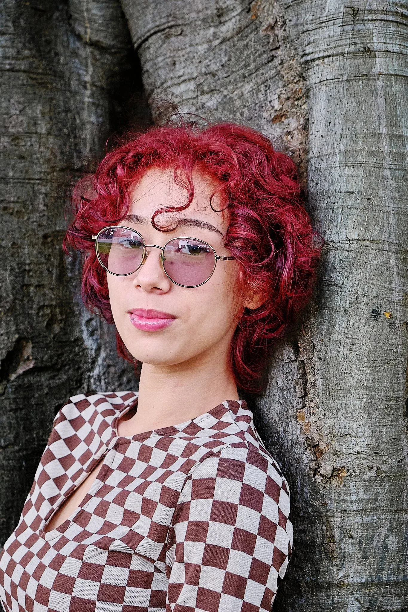 Woman with short red curly hair in checkered shirt leaning against tree trunk outdoors.