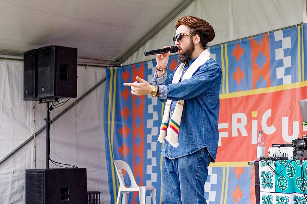 Bearded man in denim suit speaking into microphone on decorated festival stage.