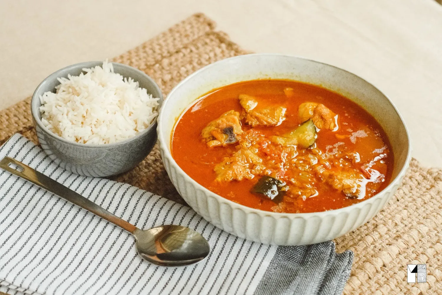 Bowl of rich tomato-based fish curry stew served with a side of white rice and a spoon.