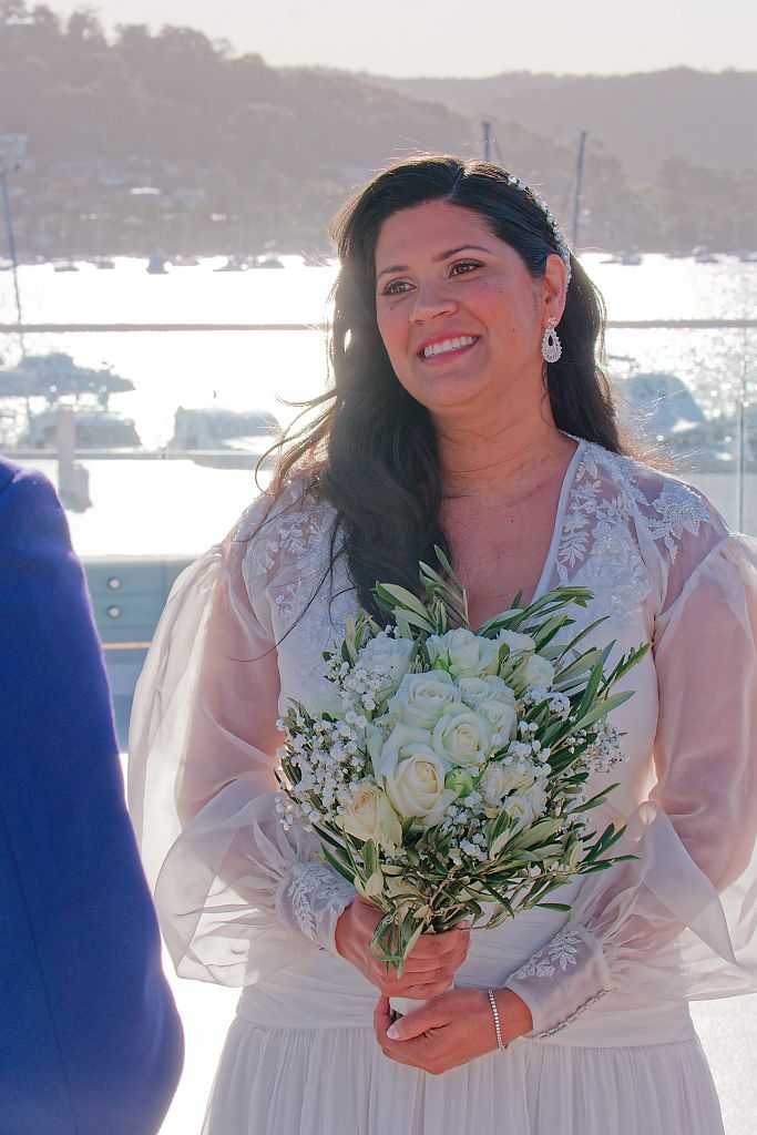 Smiling bride in a lace-sleeved white dress holds a bouquet of white roses and greenery at an outdoor waterfront wedding ceremony.