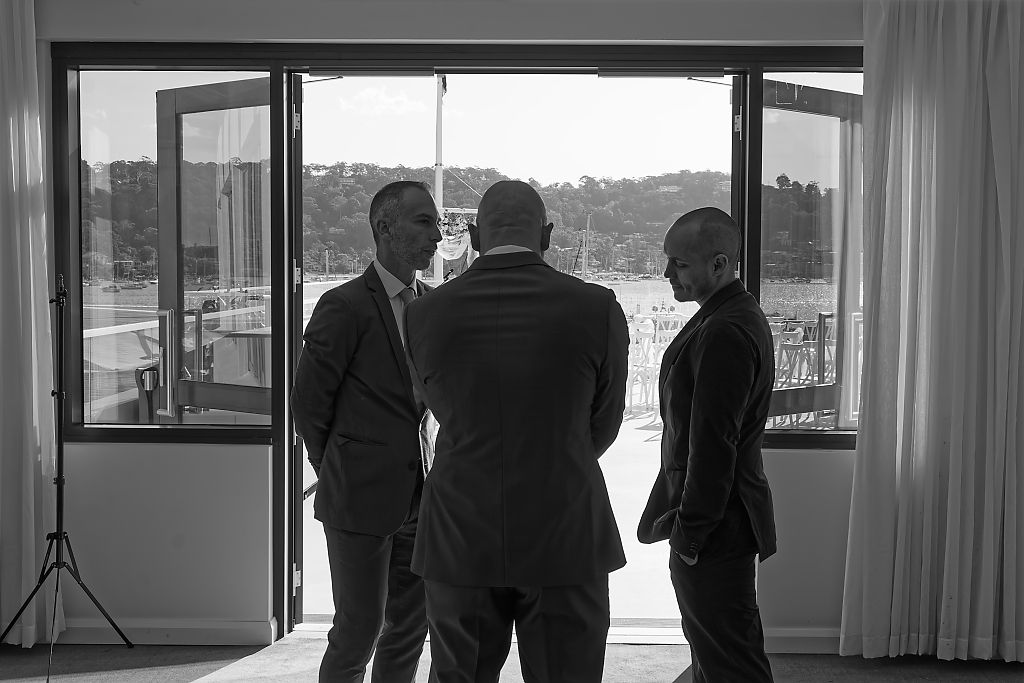 Three men in suits stand in a huddle in front of a large open window, silhouetted against bright daylight in this black and white photo.