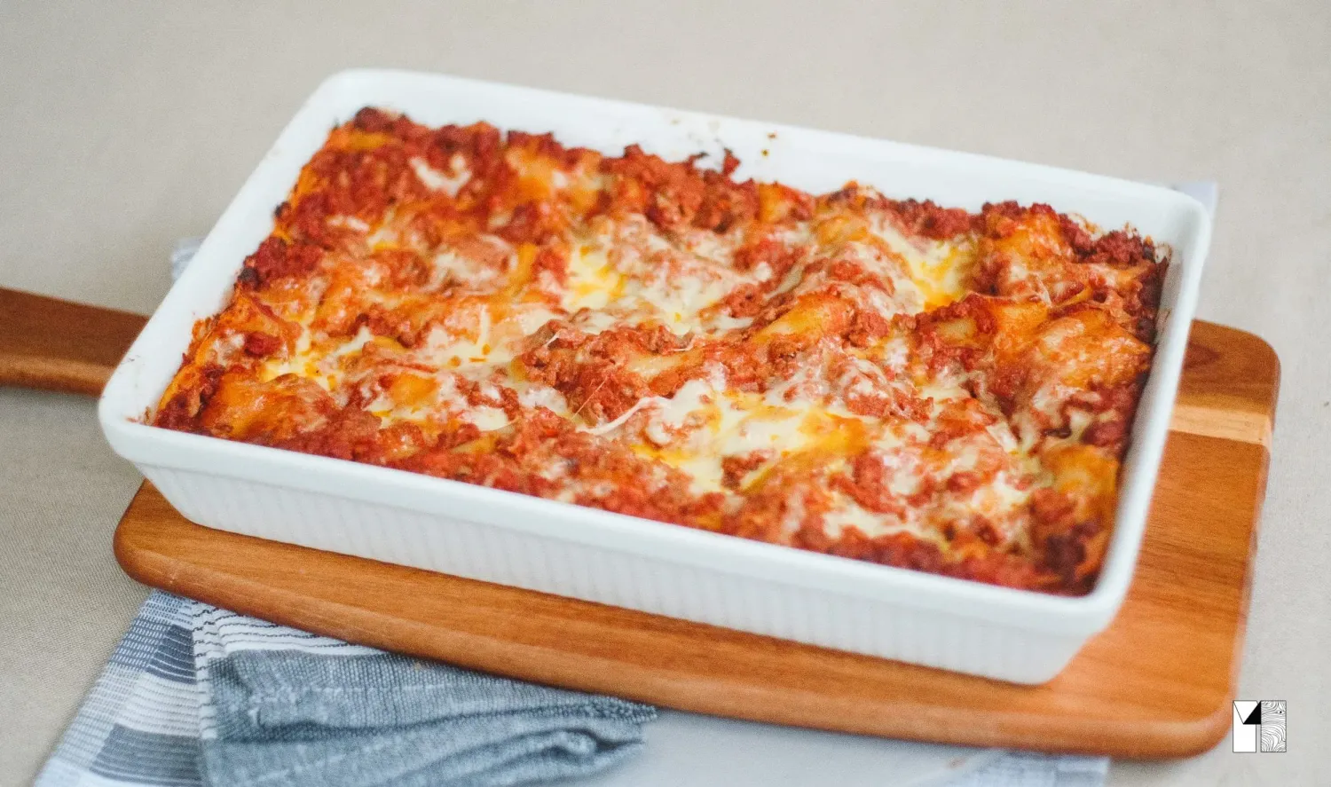 Cheesy baked lasagna with tomato sauce in a white rectangular baking dish on a wooden board.