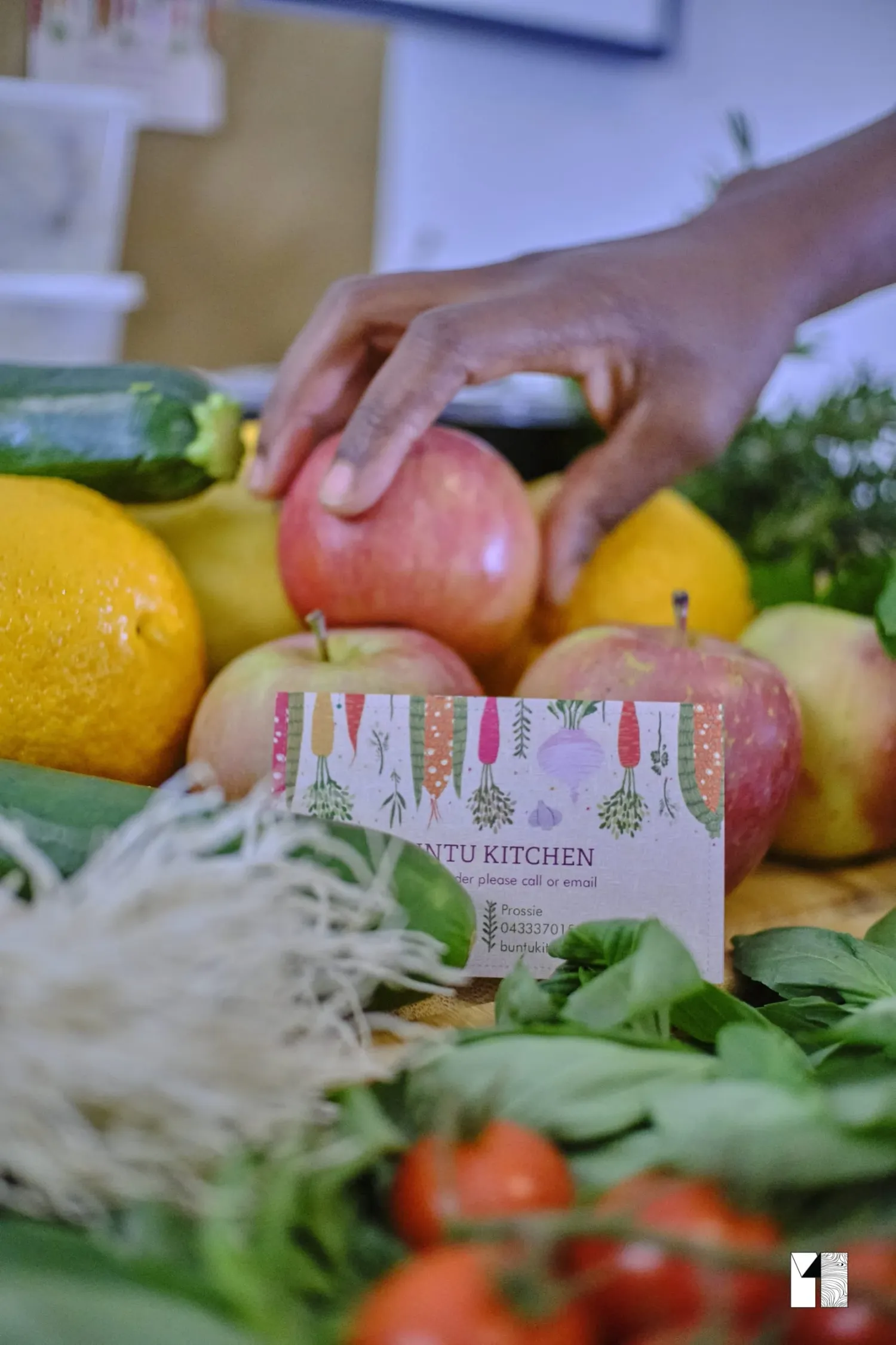 Hand picking a fresh apple from a colorful display of fruits and vegetables with Buntu Kitchen card.