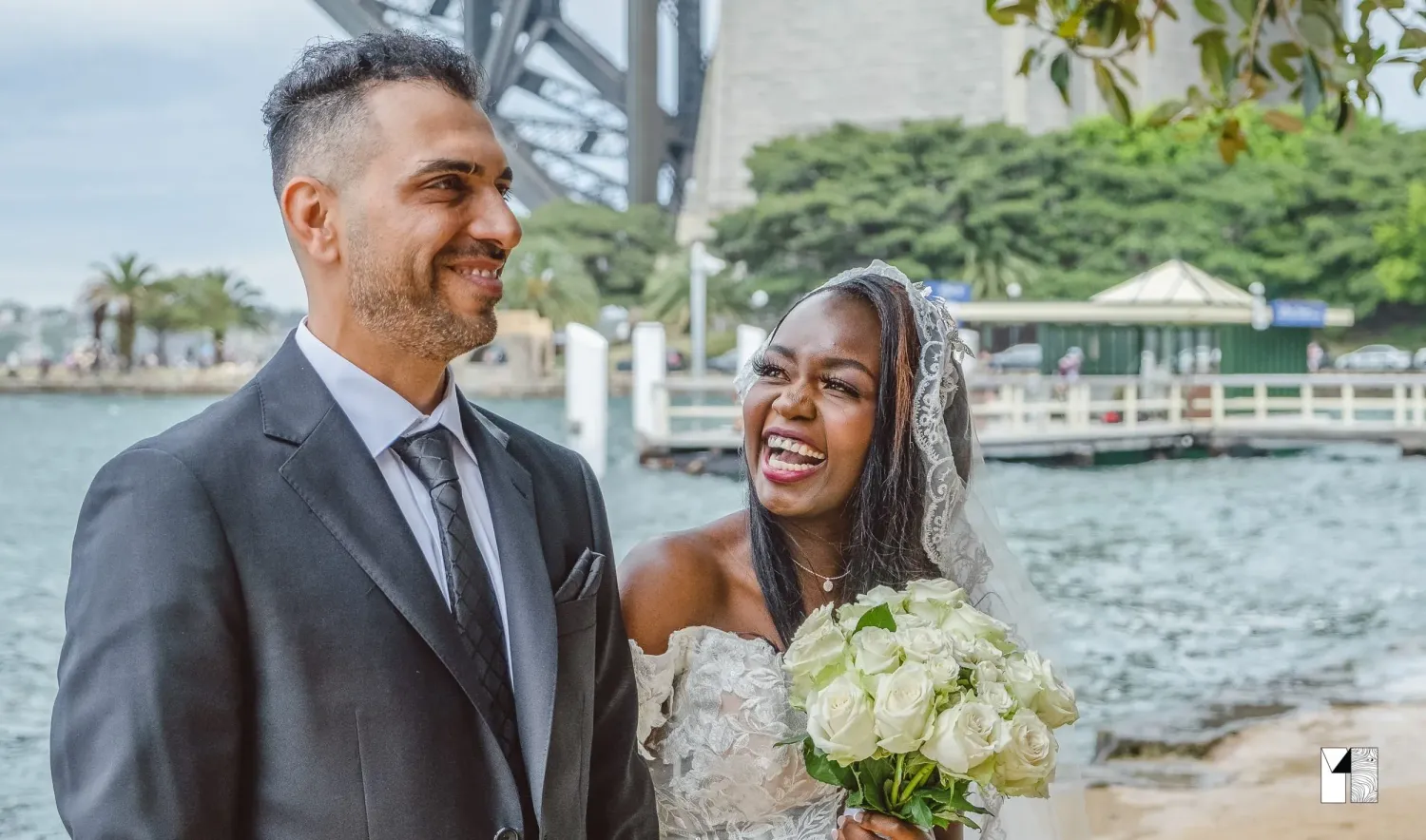 Happy interracial newlywed couple smiling together near Sydney Harbour Bridge after wedding.
