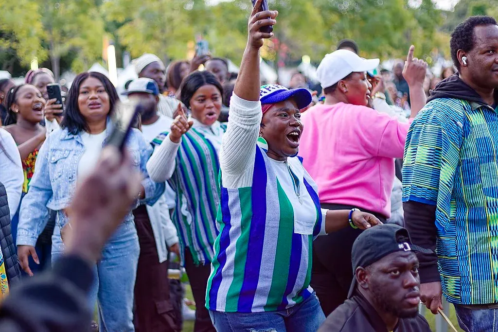Excited crowd celebrating at outdoor African cultural festival, woman in Sierra Leone flag shirt.