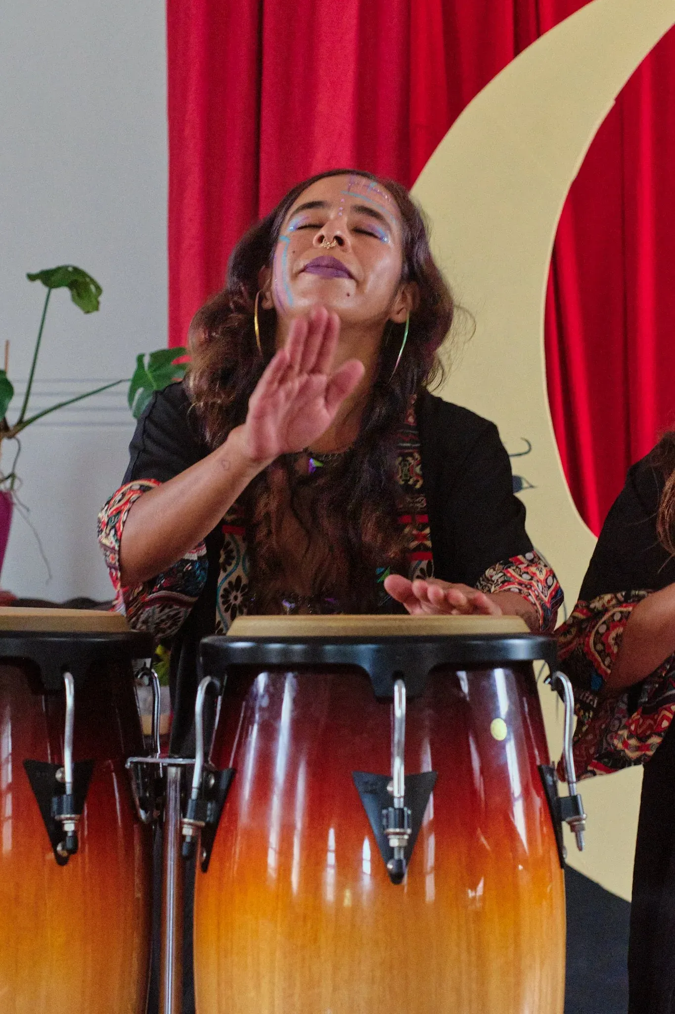 Woman playing conga drums with eyes closed and hand raised during live music performance on stage.