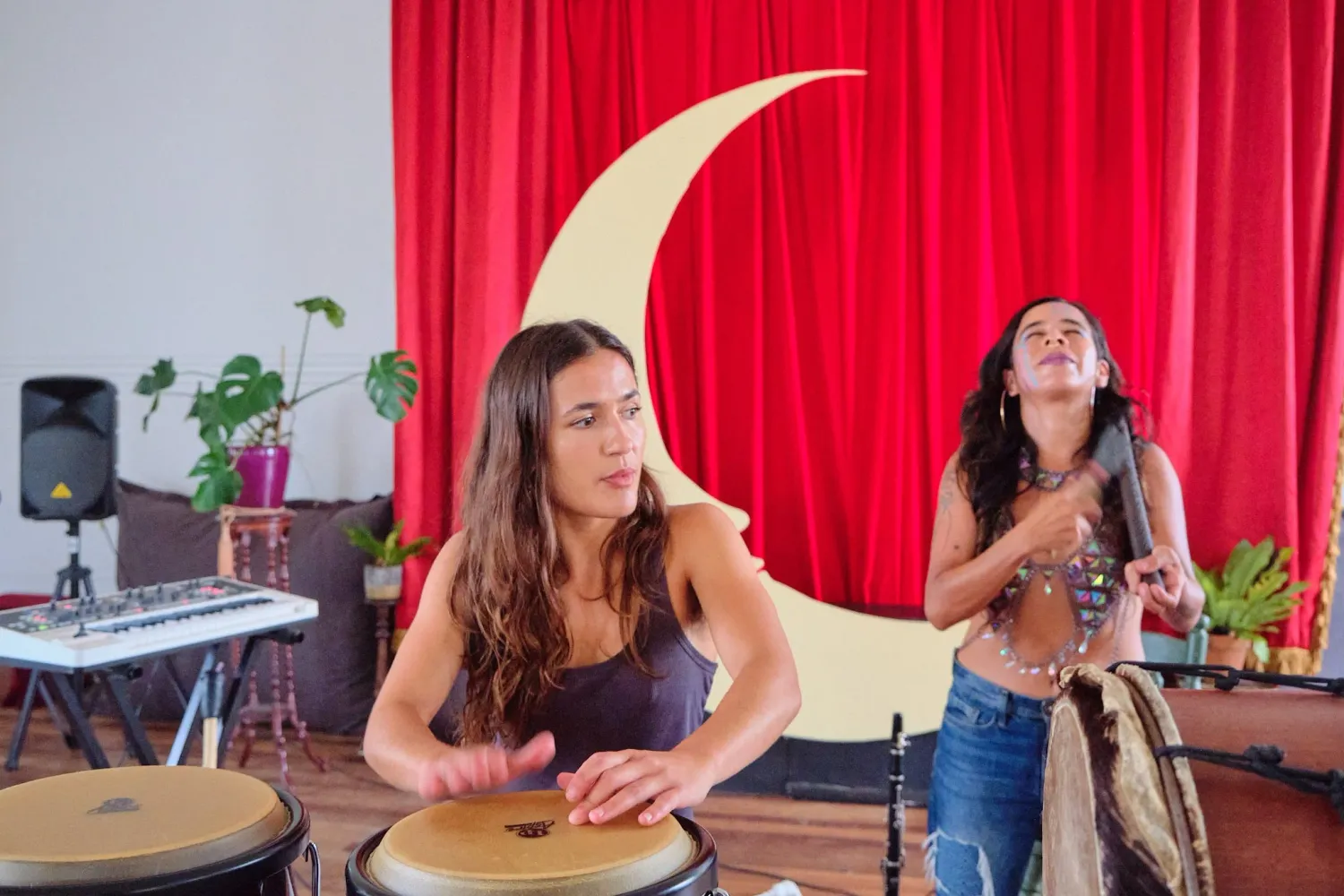 Two female musicians performing with conga drums before a red curtain and crescent moon decoration.